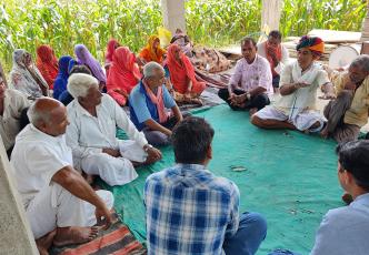 A group of adults sitting on the floor in a semi-circle under a shelter, engaging in a discussion. Some are wearing traditional Indian attire.