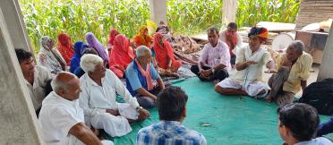 A group of adults sitting on the floor in a semi-circle under a shelter, engaging in a discussion. Some are wearing traditional Indian attire.