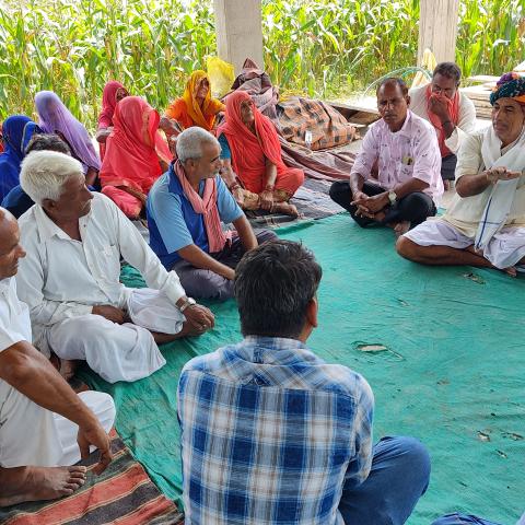 A group of adults sitting on the floor in a semi-circle under a shelter, engaging in a discussion. Some are wearing traditional Indian attire.